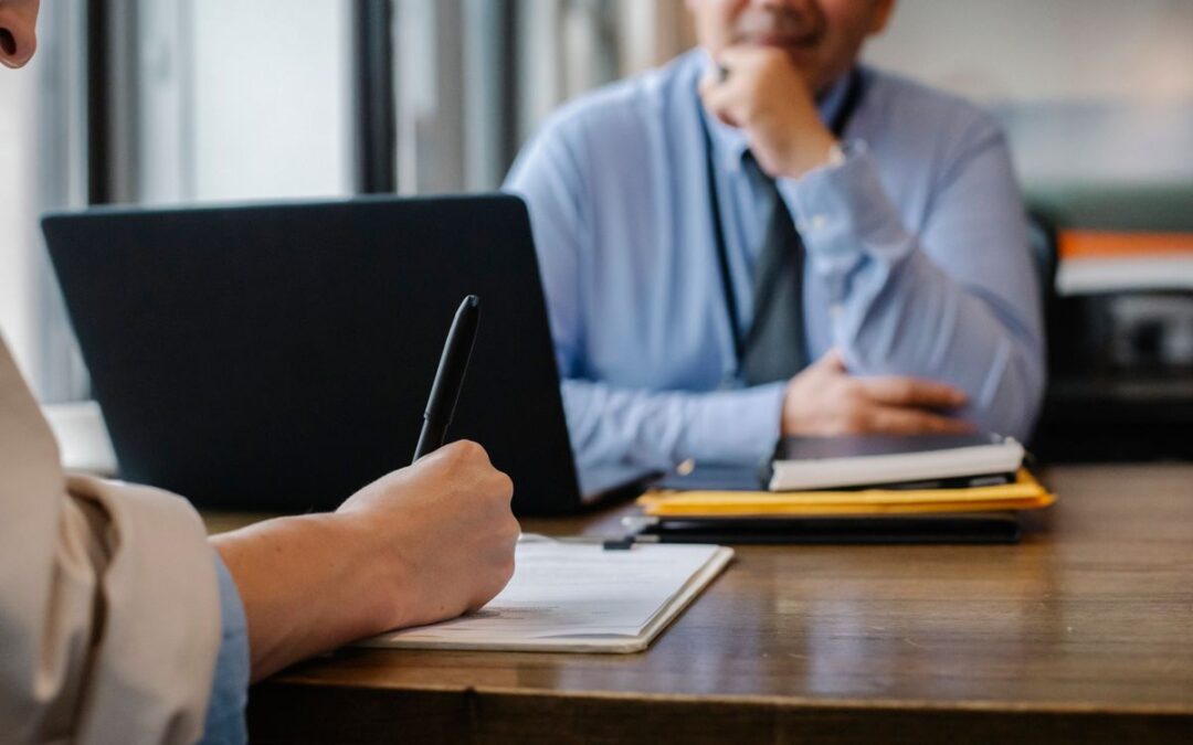 Twee zakelijke gesprekspartners aan een tafel, een van hen noteert afspraken terwijl de ander meekijkt op een laptop