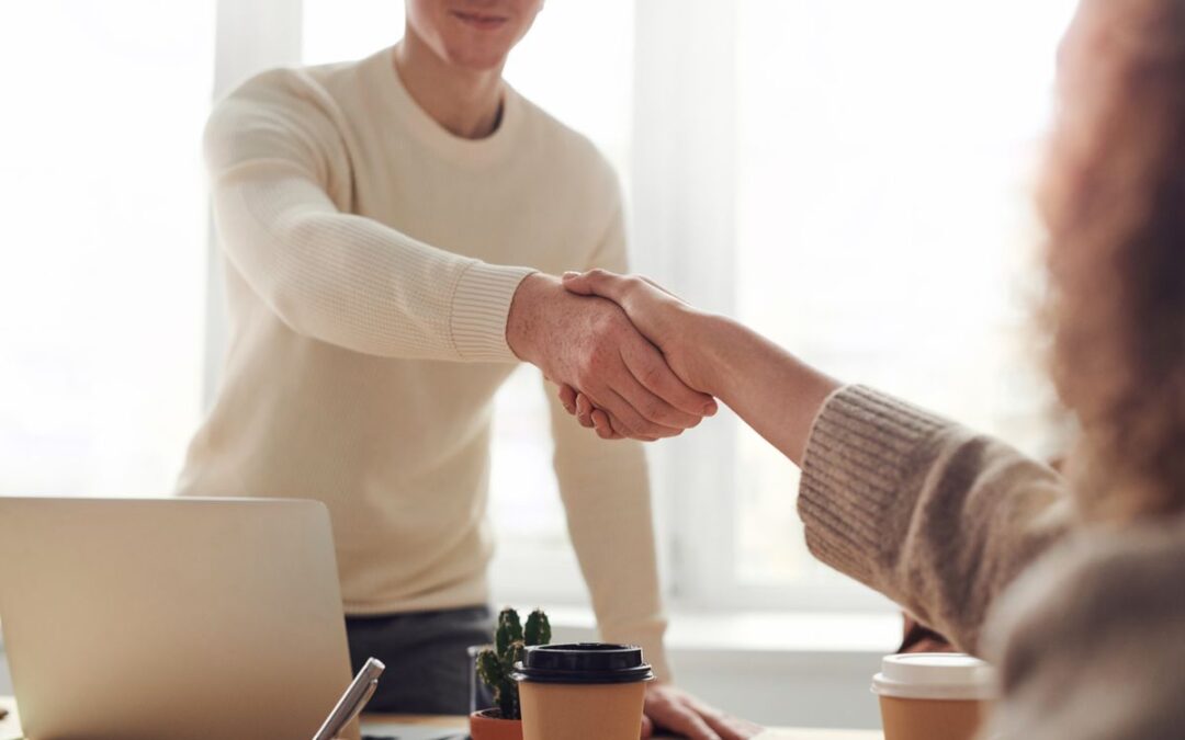Twee mensen schudden elkaar de hand boven een tafel met laptop en koffie, een eerste zakelijk contact