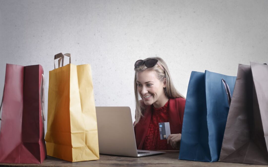 Lachende vrouw rekent online af met haar bankpas achter een laptop, omringd door kleurrijke boodschappentassen