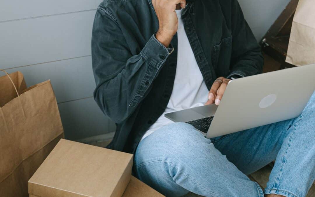 Man zit op de grond tussen pakketten en gebruikt laptop en telefoon voor een online bestelling