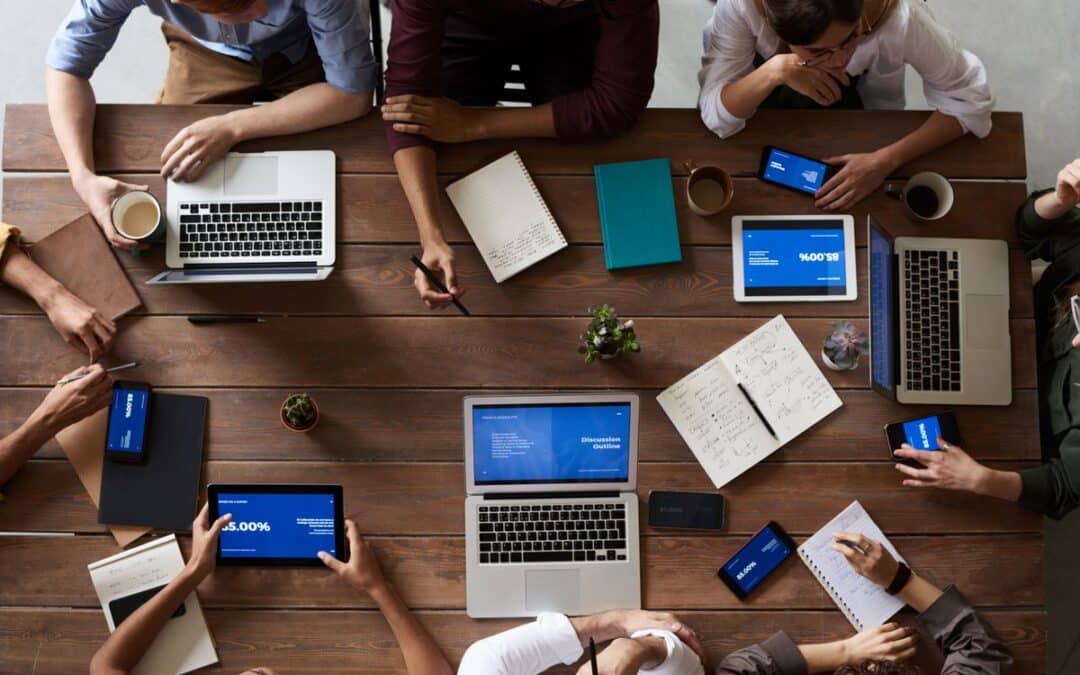 Team zit rond een houten tafel met laptops, tablets en notitieblokken tijdens een overleg
